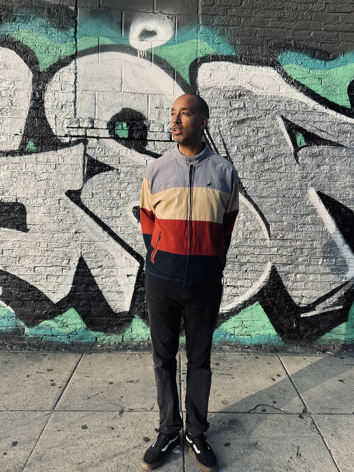 Pigeon John standing in front of a black, white, and teal graffiti wall